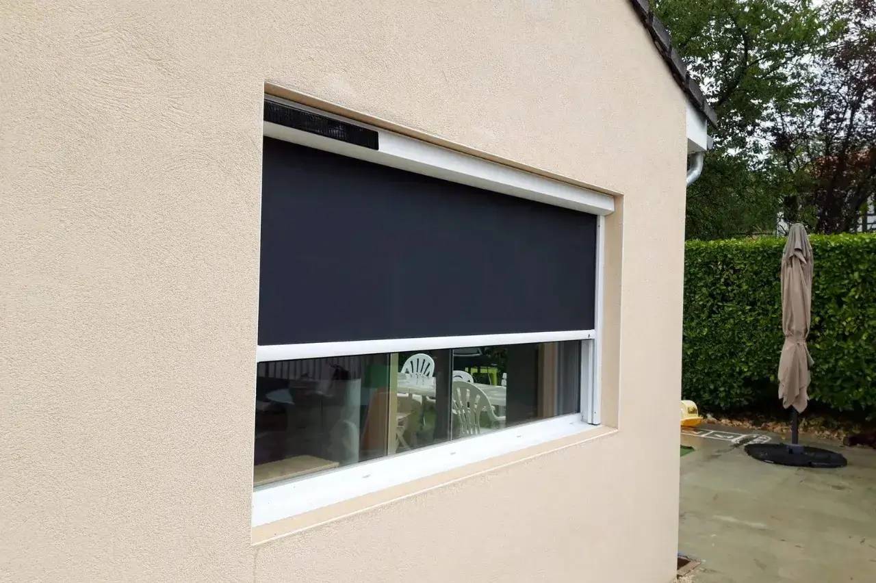 Une fenêtre extérieure avec un volet roulant sombre partiellement baissé sur un mur beige.