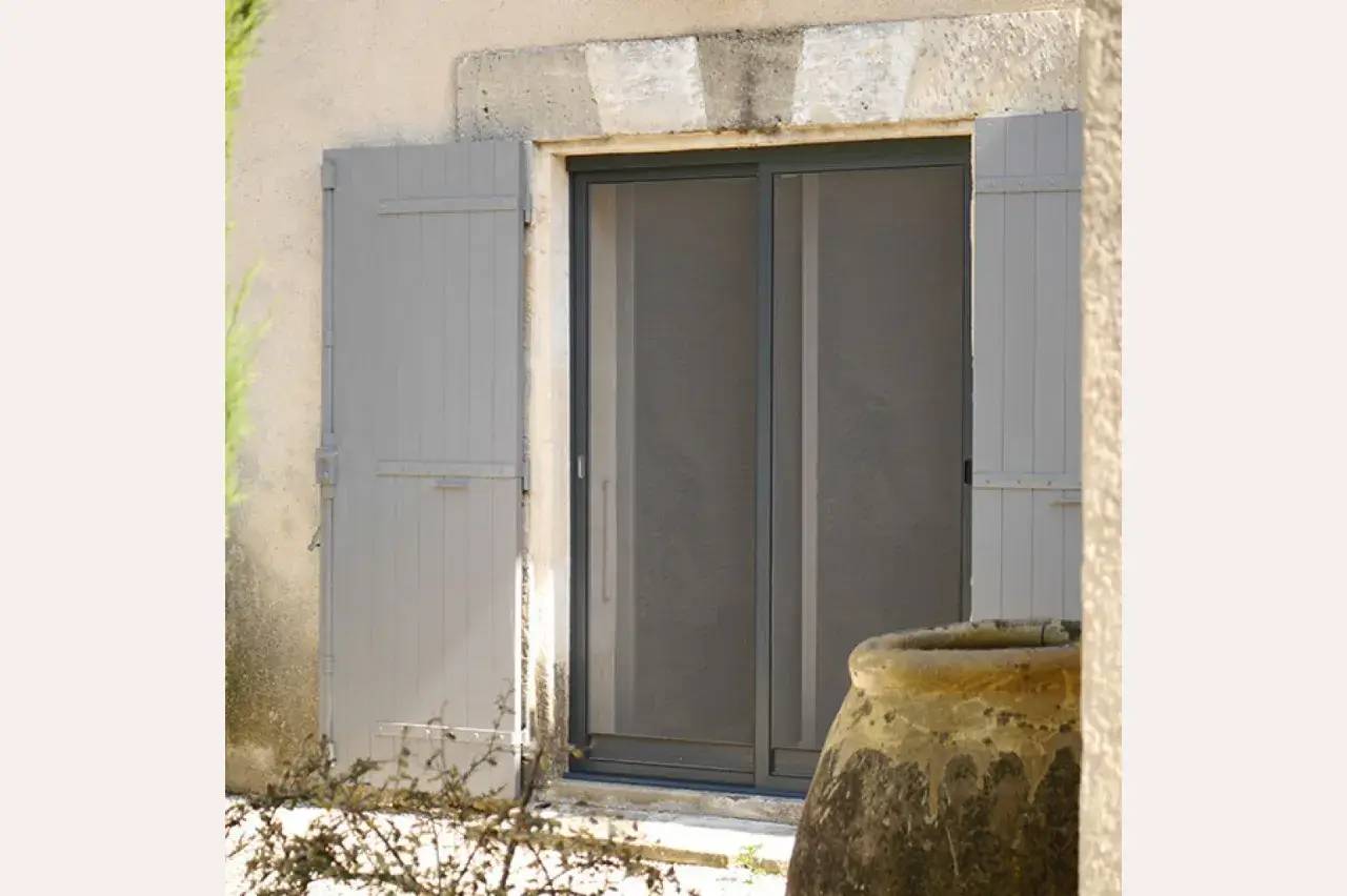 Une porte vitrée avec des volets gris est encadrée par un mur en pierre, avec une grande jarre en terre cuite au premier