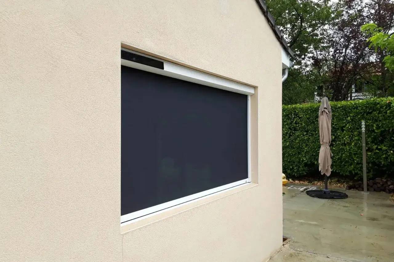 Un auvent beige équipé d’un store enrouleur noir sur une terrasse avec des haies vertes et un parasol fermé.