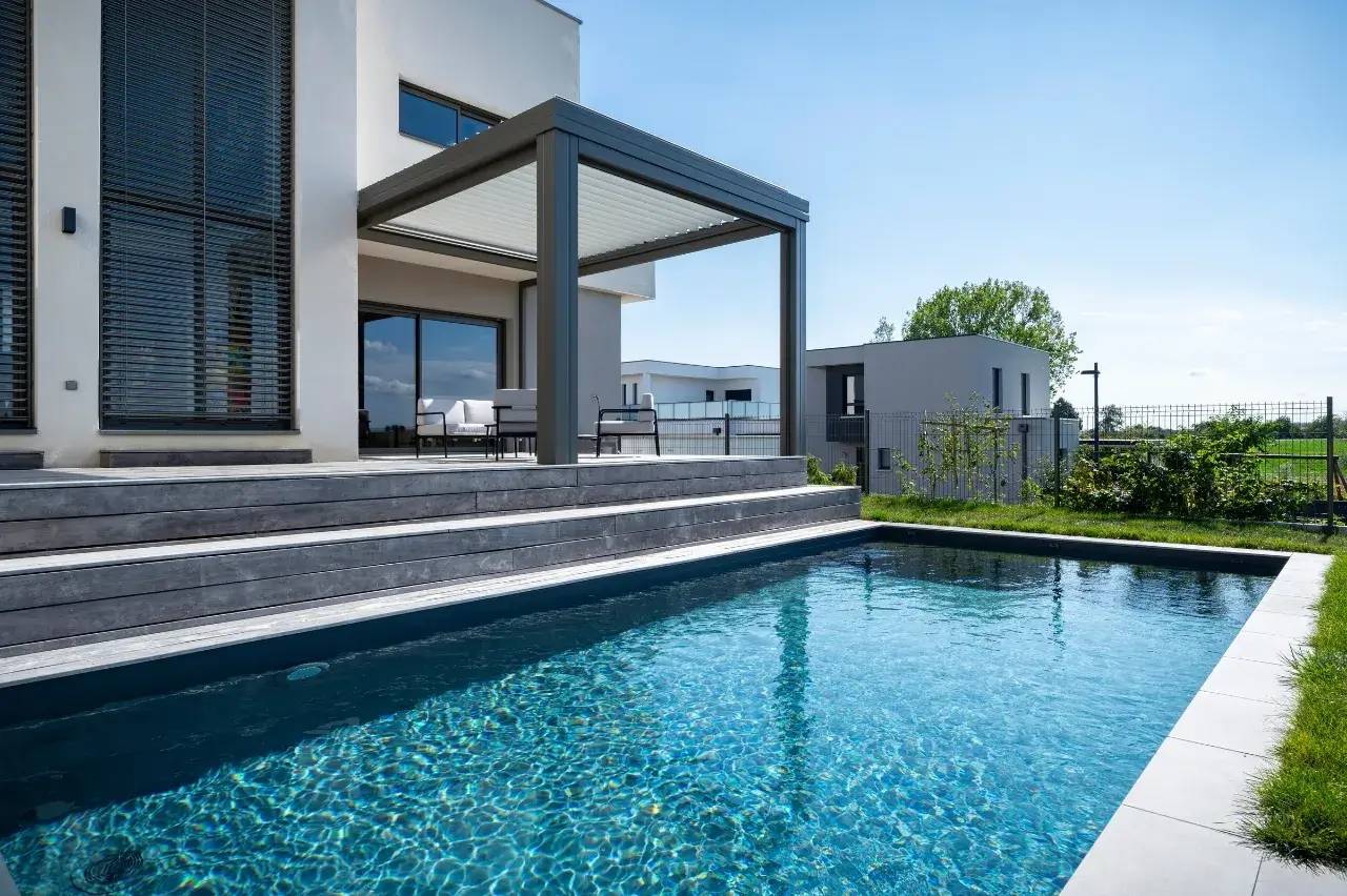 Une maison moderne avec une terrasse couverte et une piscine étincelante sous un ciel dégagé.