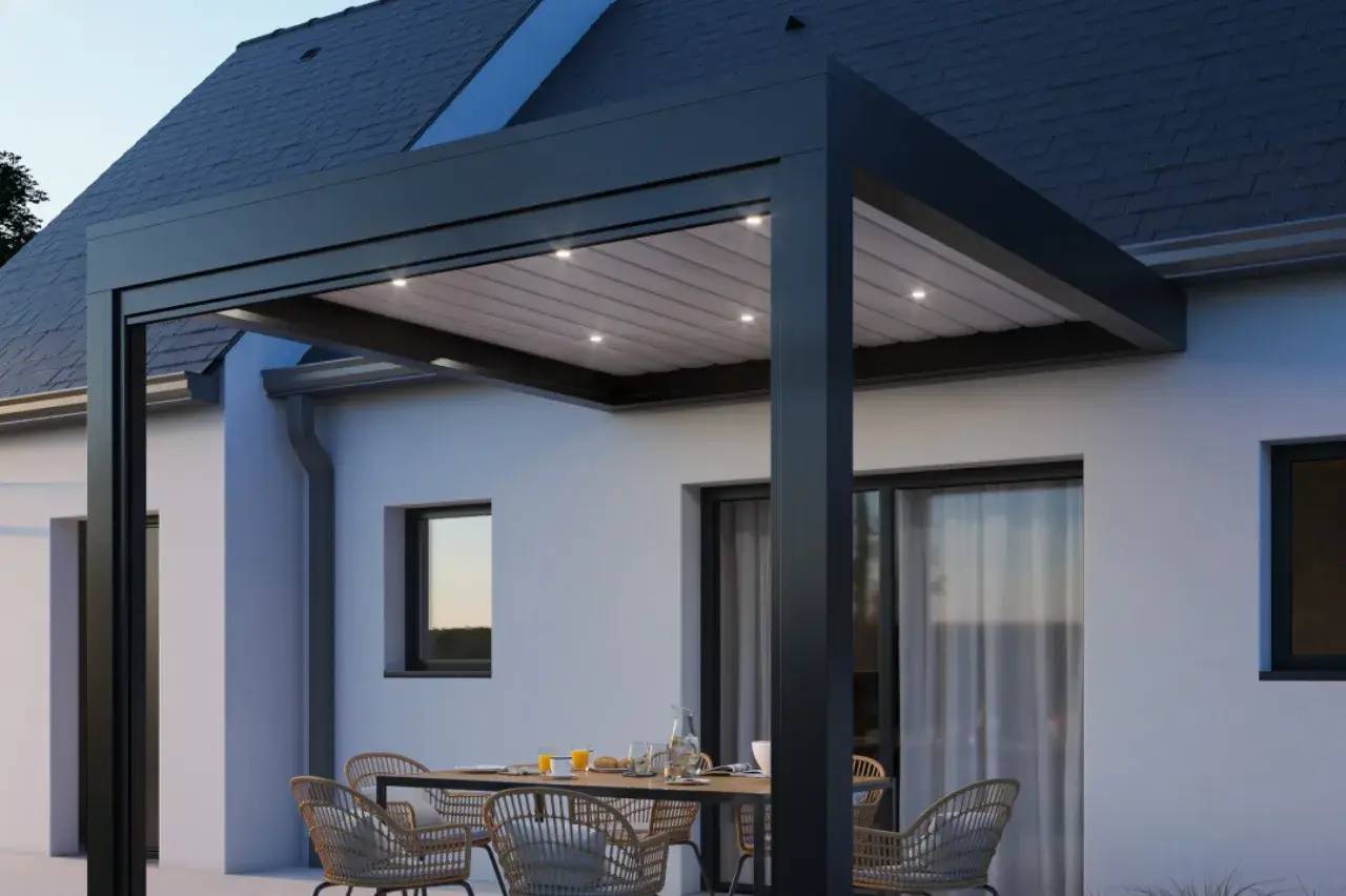 Une terrasse moderne avec une pergola noire éclairée, équipée d'une table et de chaises en rotin, est attenante à une maison blanche