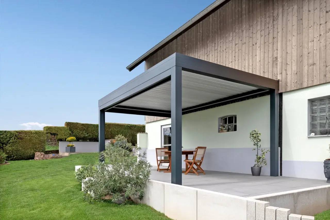Une terrasse moderne avec une pergola en métal gris, meublée de chaises et d'une table en bois, donnant sur un jardin verdoyant