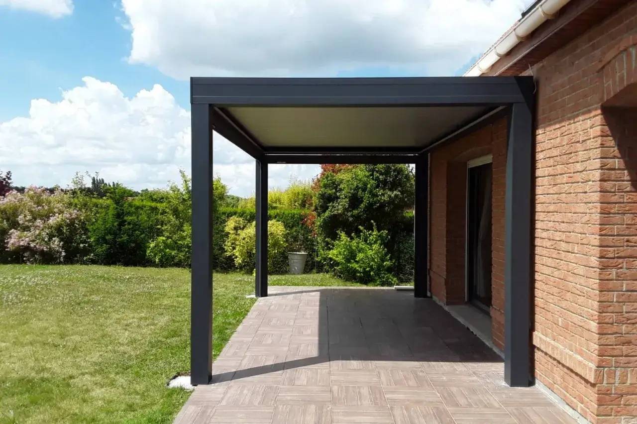 Une élégante pergola en métal sombre prolonge une terrasse, encadrée de verdure luxuriante et d'un mur en briques