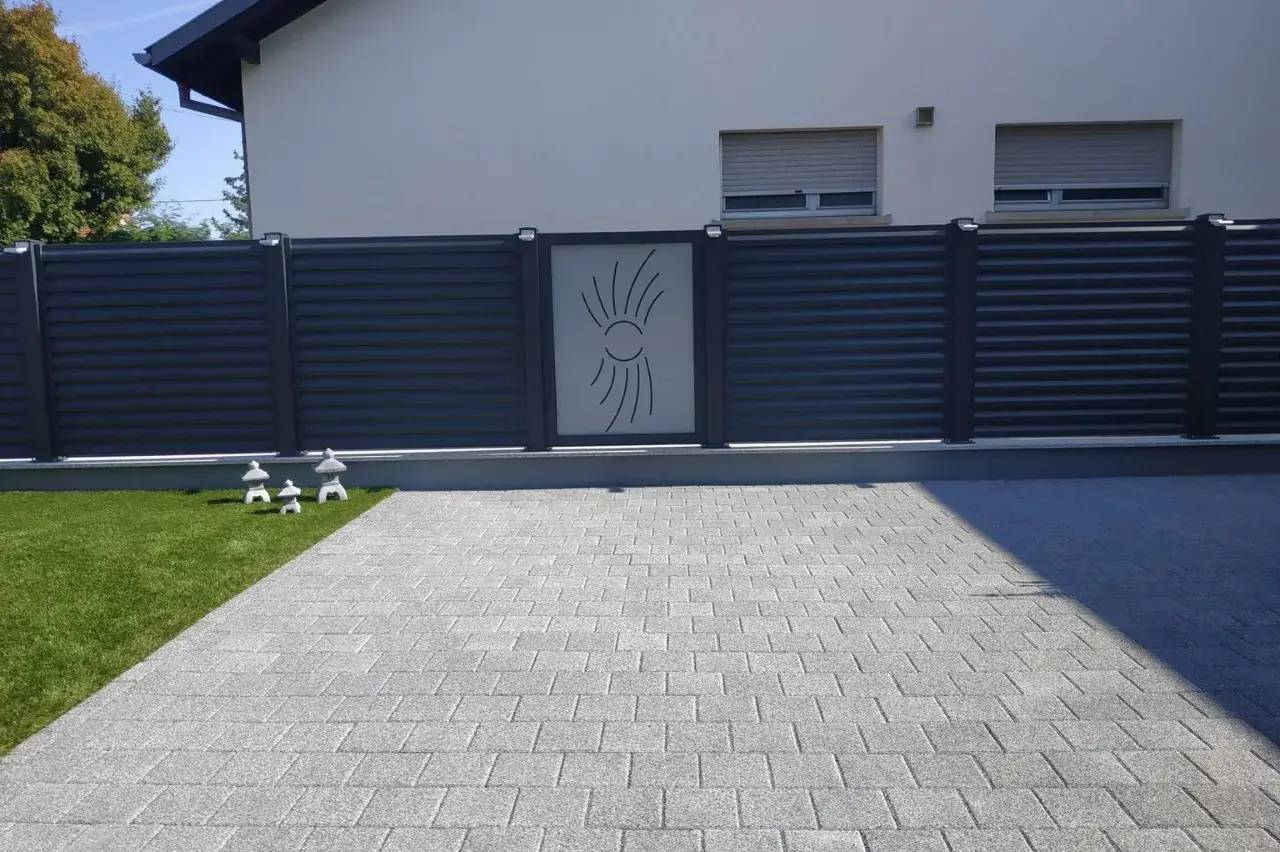 Une clôture moderne en métal gris avec un motif central stylisé est entourée de verdure et d'un sol pavé.