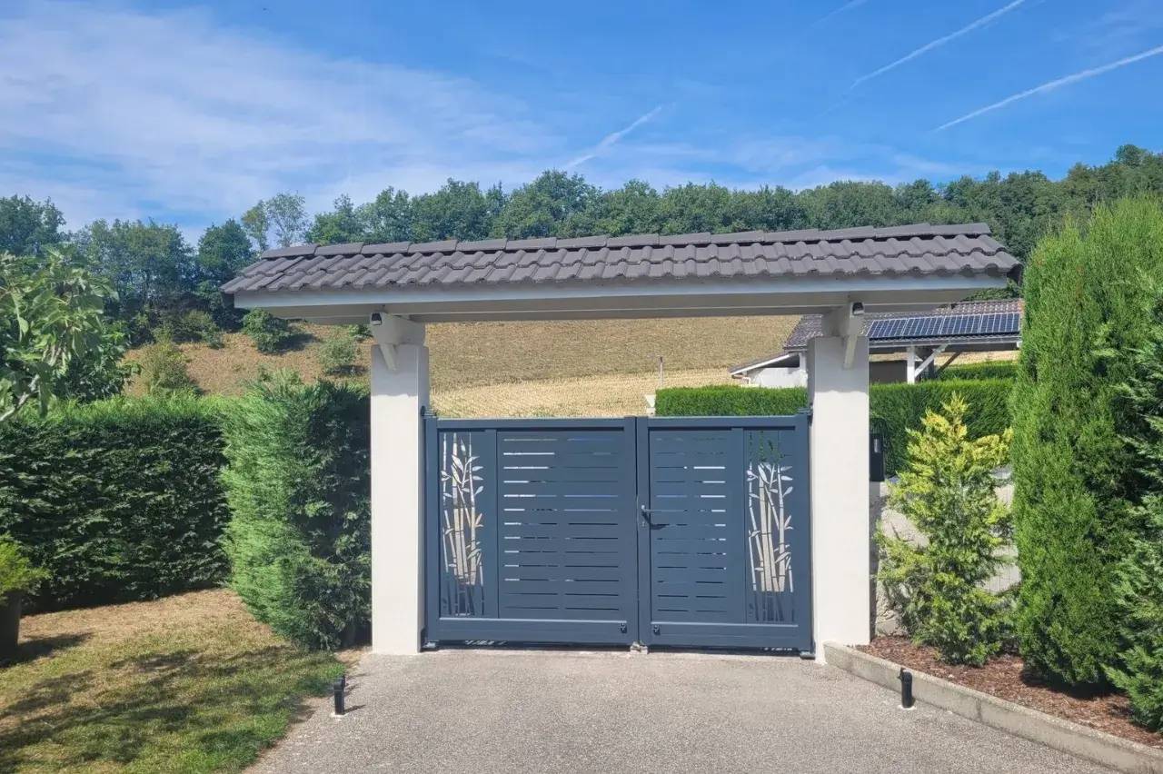 Portail moderne en métal gris foncé avec motifs de bambou, encadré de colonnes blanches, entouré de verdure sous un