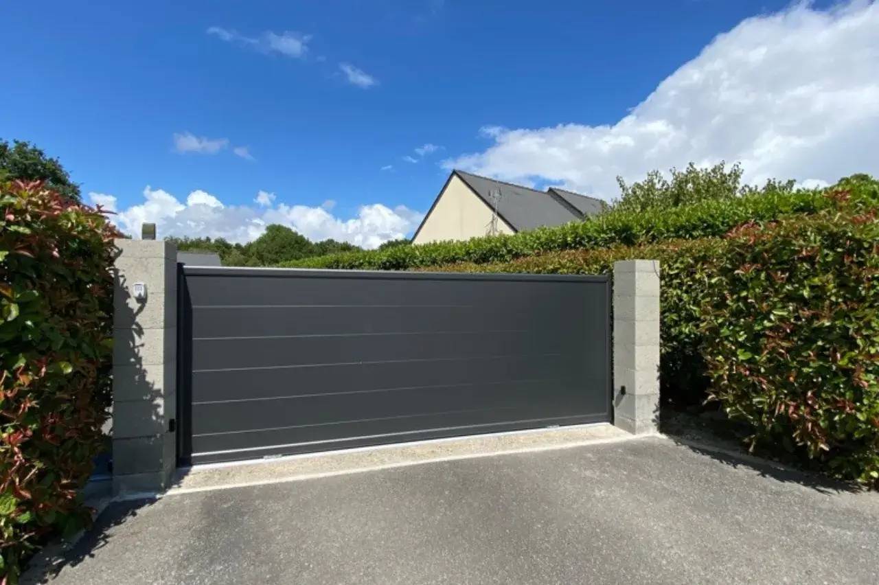 Portail moderne gris anthracite entouré de haies verdoyantes, sous un ciel bleu parsemé de quelques nuages.