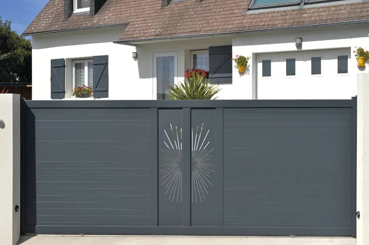 Portail moderne gris anthracite avec motif géométrique central devant une maison blanche aux volets noirs.