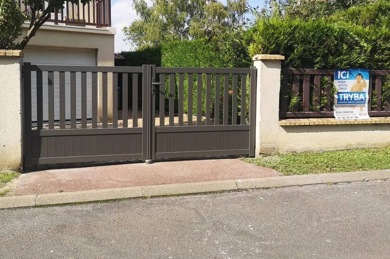 Portail moderne en métal gris devant un garage, entouré de haies et d'un panneau publicitaire.