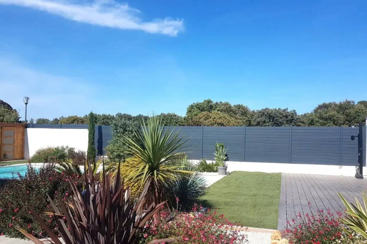 Jardin moderne avec pelouse soignée, plantes exotiques et clôture grise sous un ciel bleu clair.