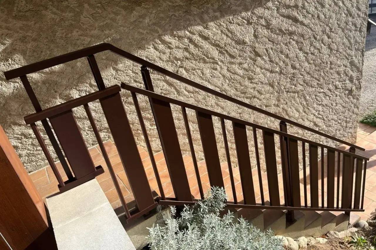 Un escalier en bois descend le long d'un mur en pierre texturée, entouré de végétation.