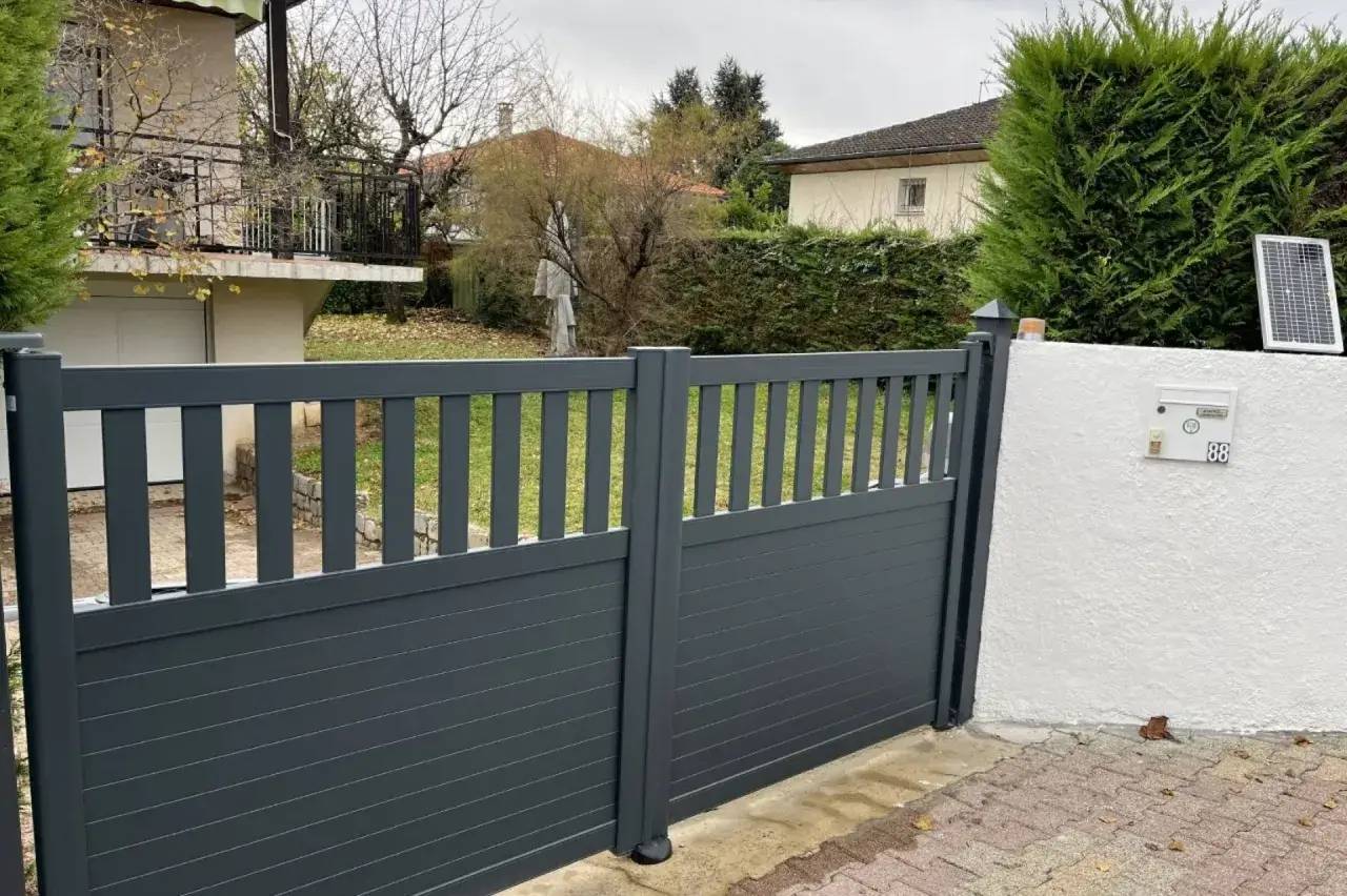 Portail moderne en aluminium gris foncé, jouxtant un mur blanc et donnant sur un jardin arboré.