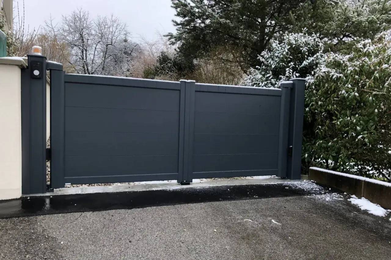 Un portail gris moderne se dresse devant un paysage hivernal avec des arbres enneigés.