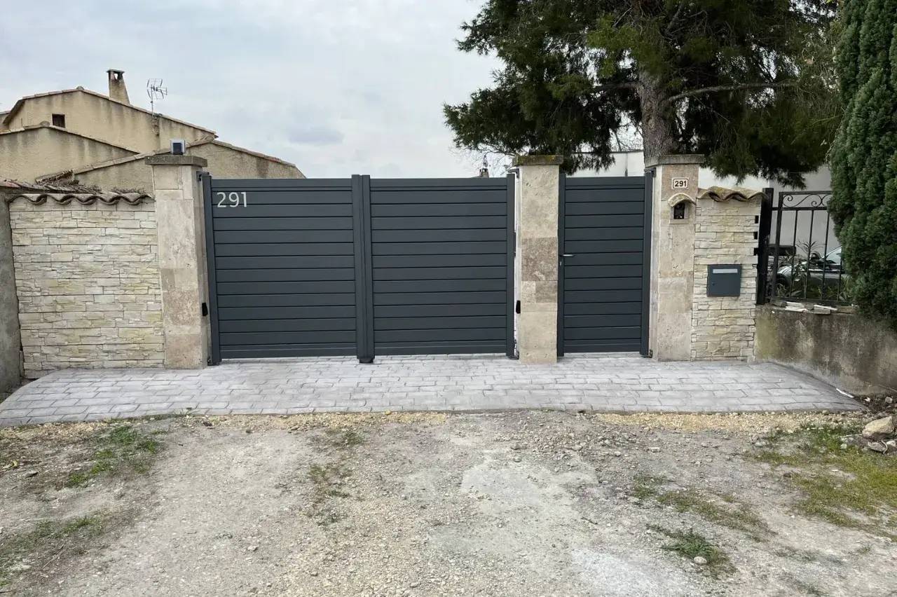 Portail moderne gris en métal avec numéros de maison visibles, entouré de murs en pierre et de végétation.