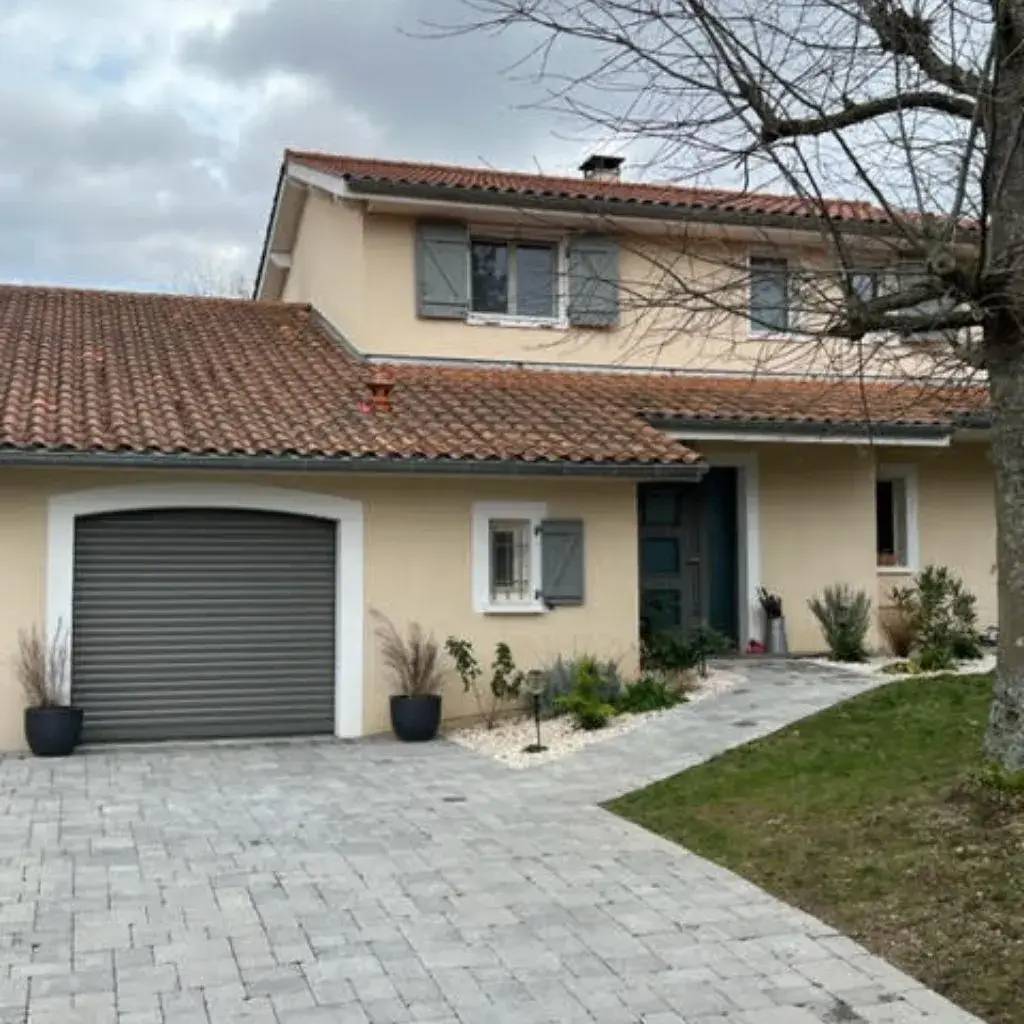 Maison moderne à deux étages avec garage, façade beige, volets gris et toit en tuiles rouges entourée d’un jardin soigné.