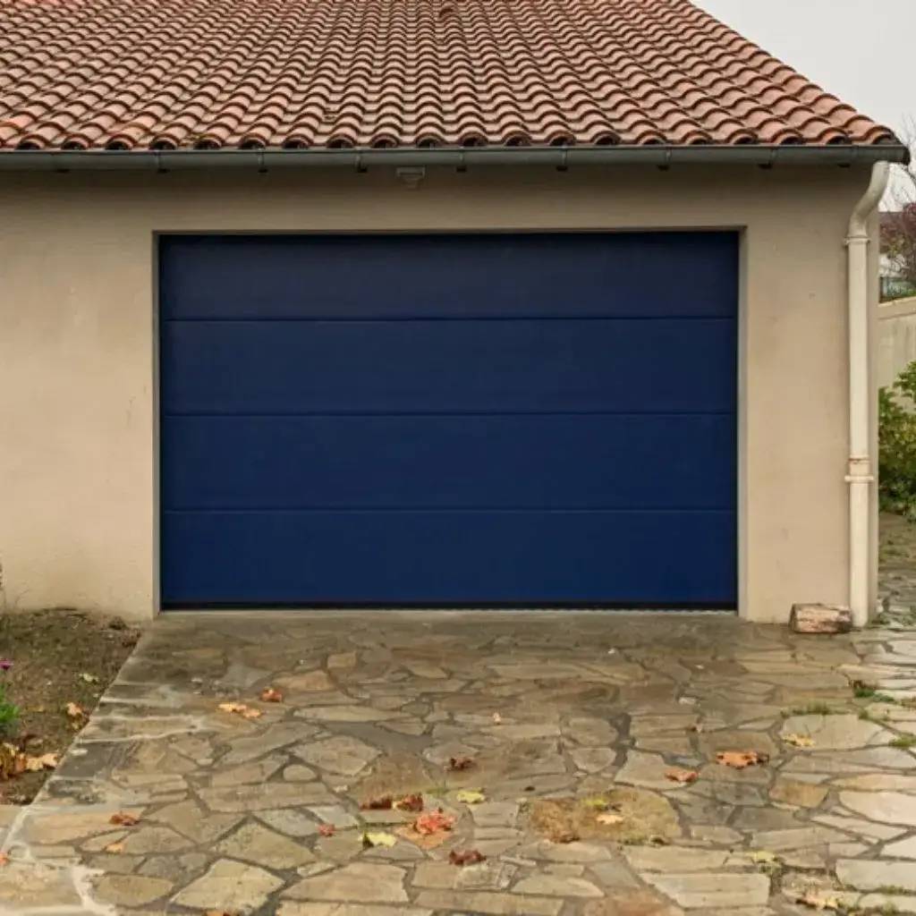 Une porte de garage bleu foncé est installée sur un mur beige avec un toit en tuiles rouges.