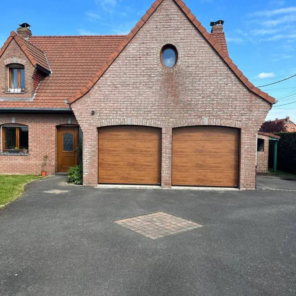 Maison en briques rouges avec un toit en tuiles, deux portes de garage en bois, et une entrée surmontée d'une fenêtre ronde