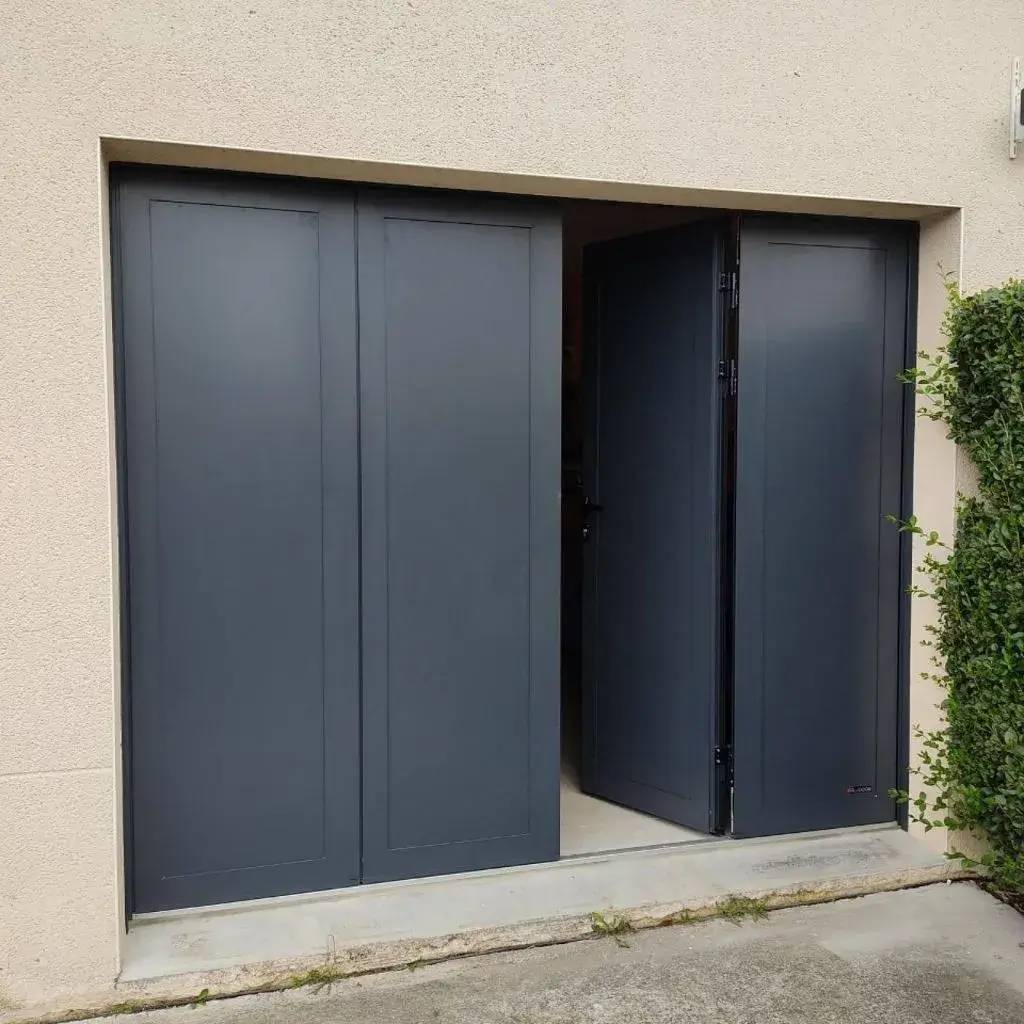 Une porte de garage moderne anthracite à deux battants partiellement ouverte, entourée d'un mur clair et de verdure.