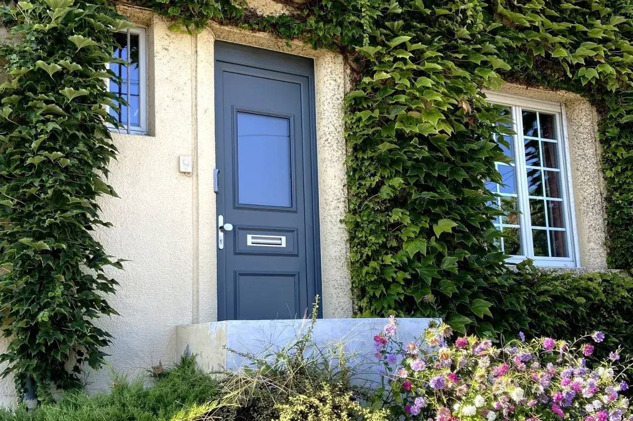 Porte d'entrée moderne entourée de lierre et de fleurs colorées sur un mur en crépi blanc.