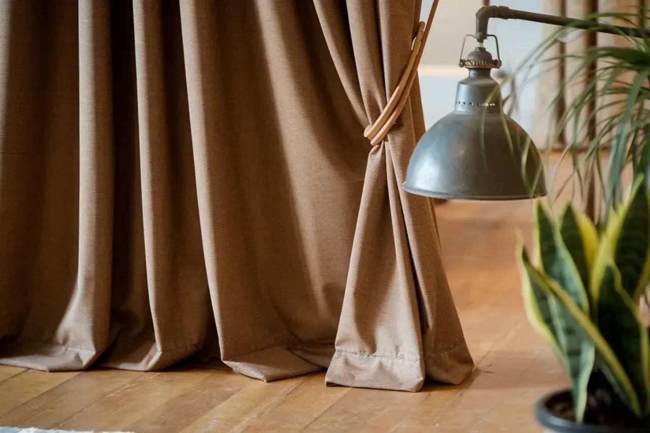 Un rideau en tissu beige tombe gracieusement sur un plancher en bois, accompagné d'une lampe suspendue et d'une plante verte.