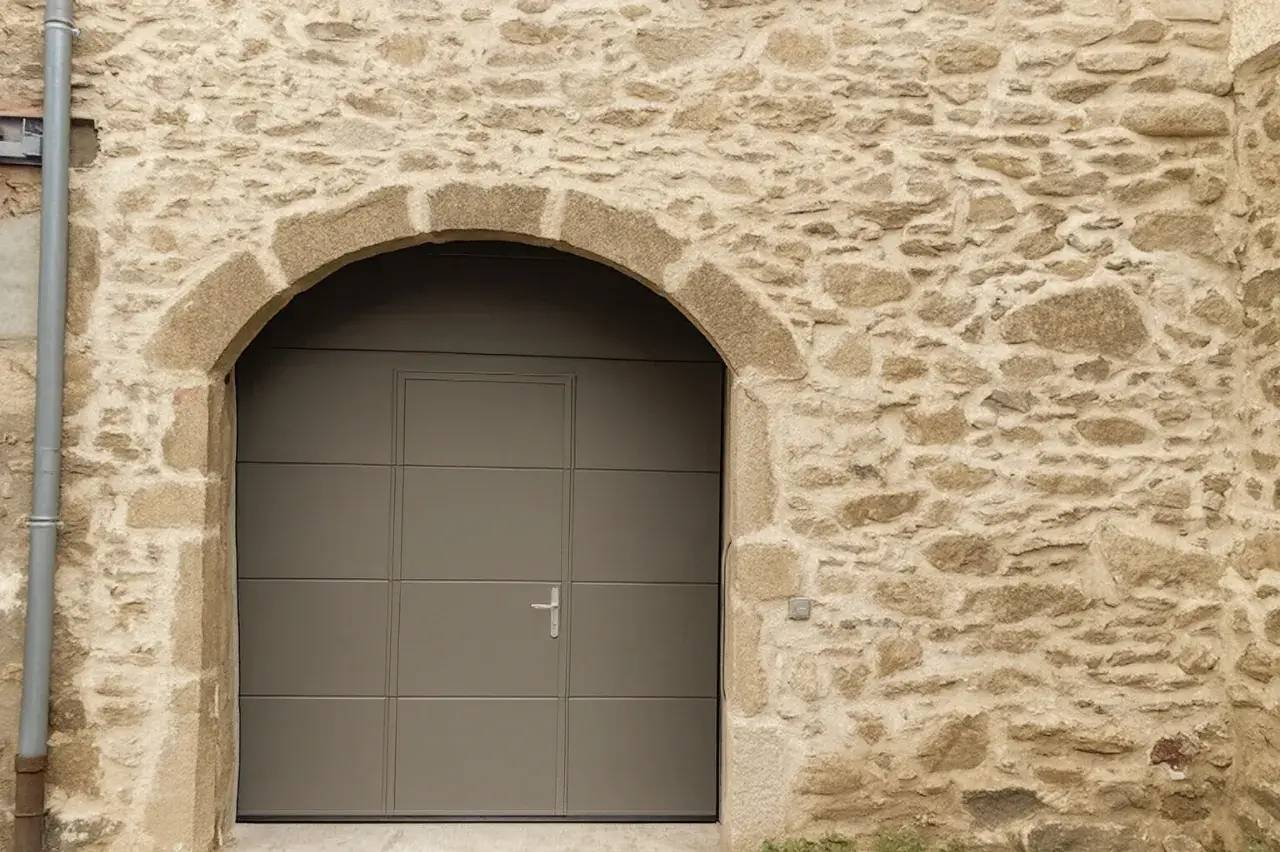 Une porte moderne de couleur grise est intégrée dans un mur en pierre à arc en plein cintre.