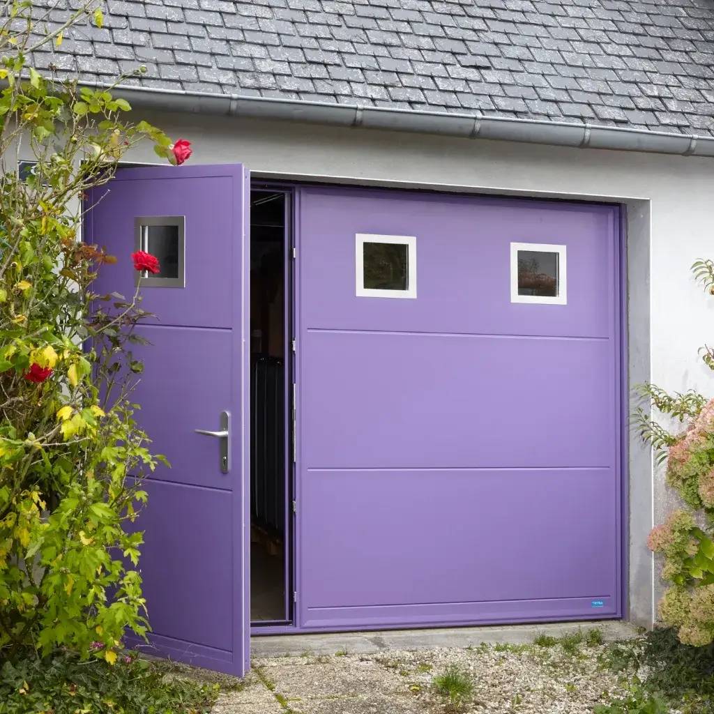 Une porte de garage violette est partiellement ouverte, encadrée par des plantes fleuries, sous un toit en tuiles grises.