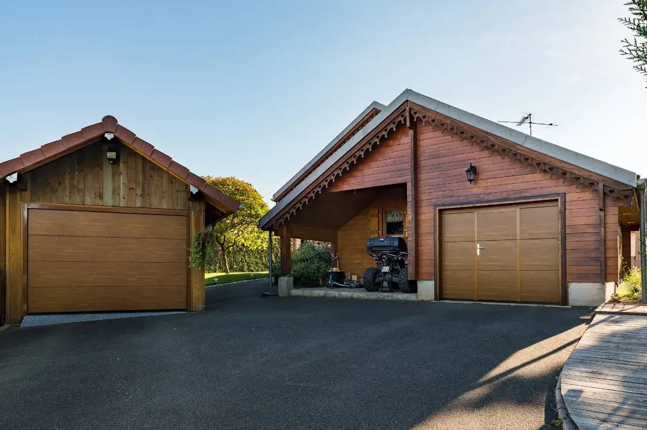 Deux garages en bois avec portes automatiques se trouvent sur une allée pavée, entourés de verdure.