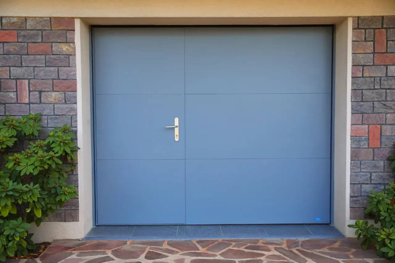 Une porte de garage bleue avec poignée, encadrée de murs en pierre et entourée de végétation.
