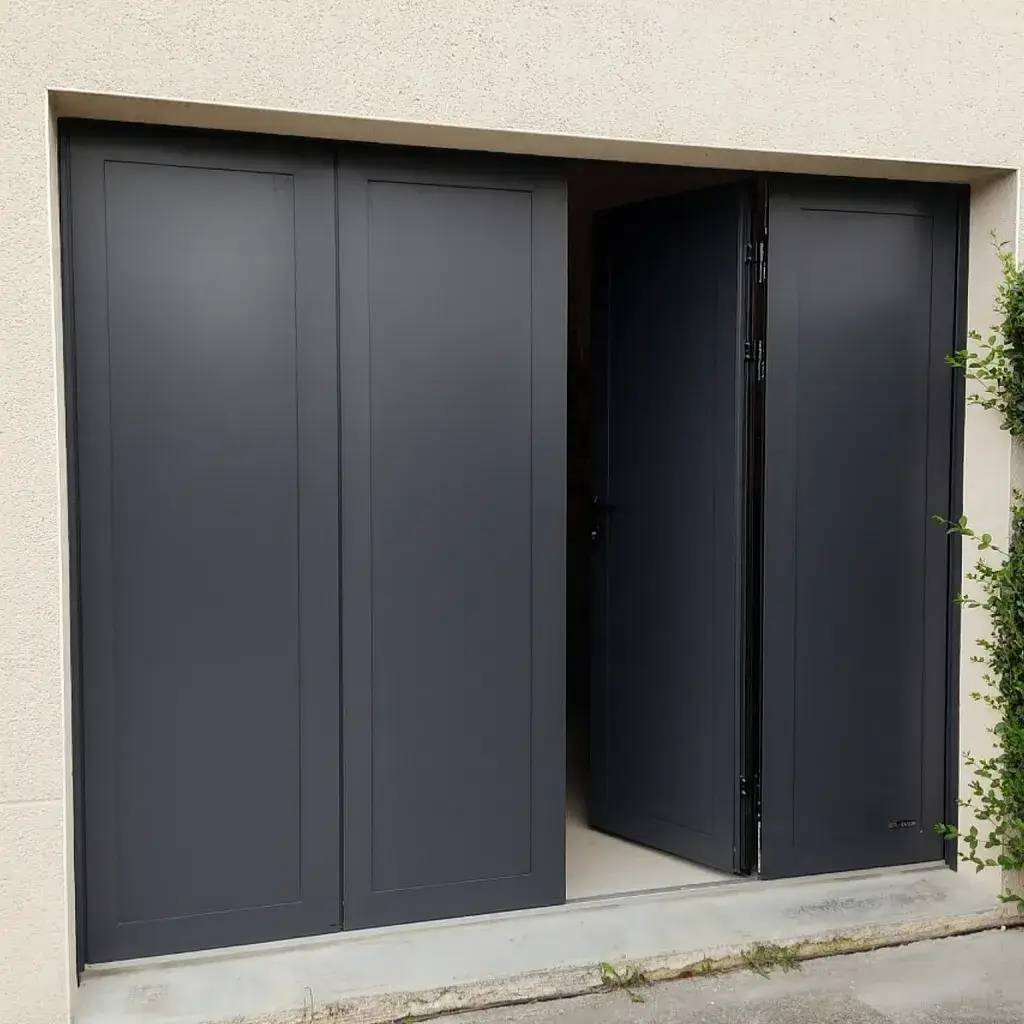 Une porte de garage moderne, gris anthracite, légèrement entrouverte sur un mur en béton clair avec quelques plantes grimpantes sur le côté.