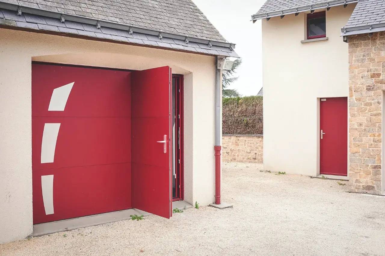Une porte de garage rouge moderne est partiellement ouverte devant une maison en pierre avec un espace extérieur pavé.