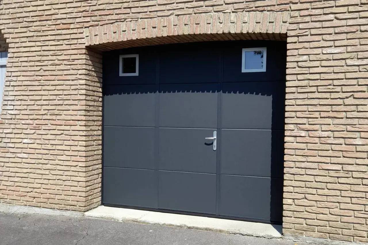Porte de garage moderne en métal noir avec poignées argentées, intégrée dans un mur de briques.