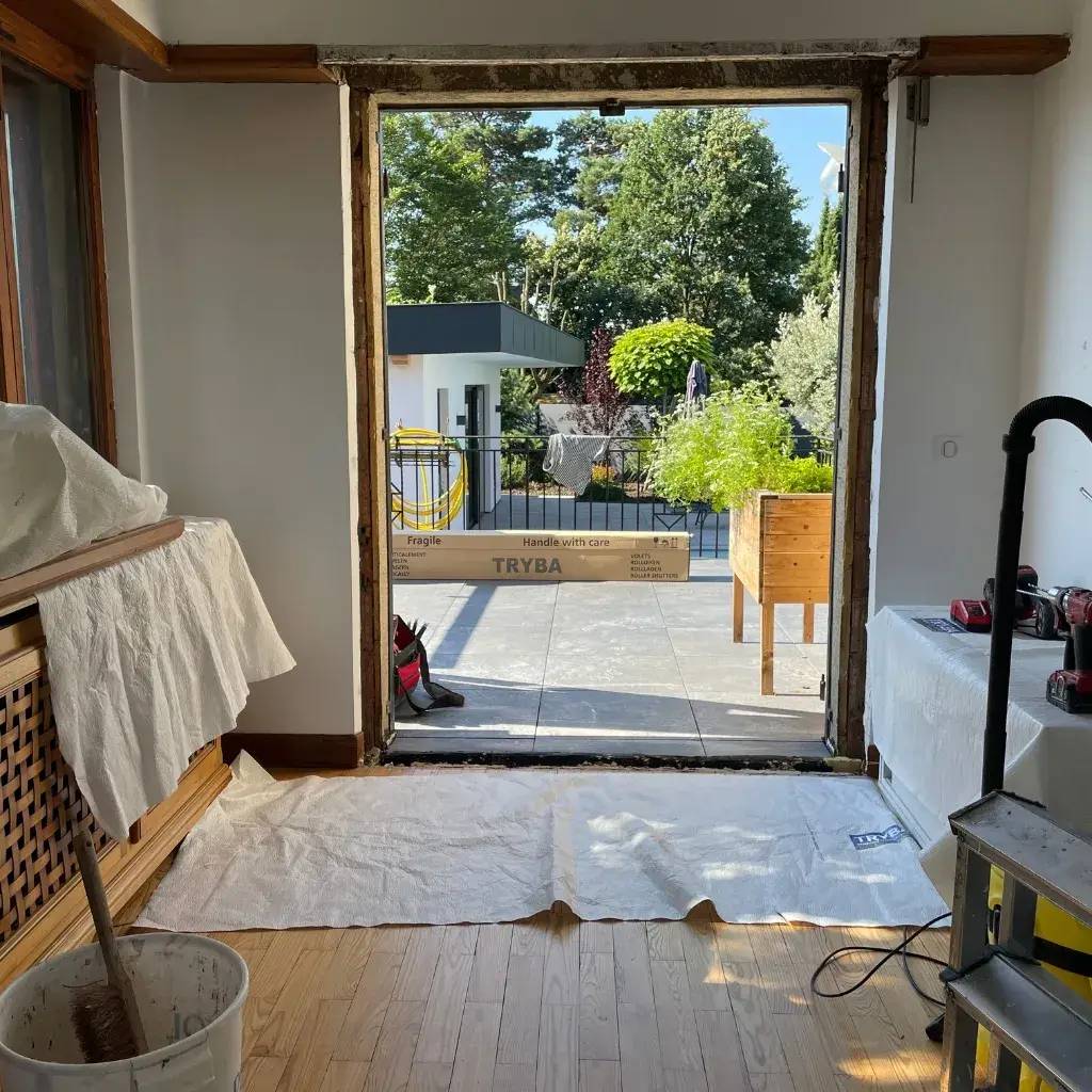 Vue d'une pièce intérieure en rénovation avec une grande ouverture donnant sur une terrasse ensoleillée entourée de verdure.