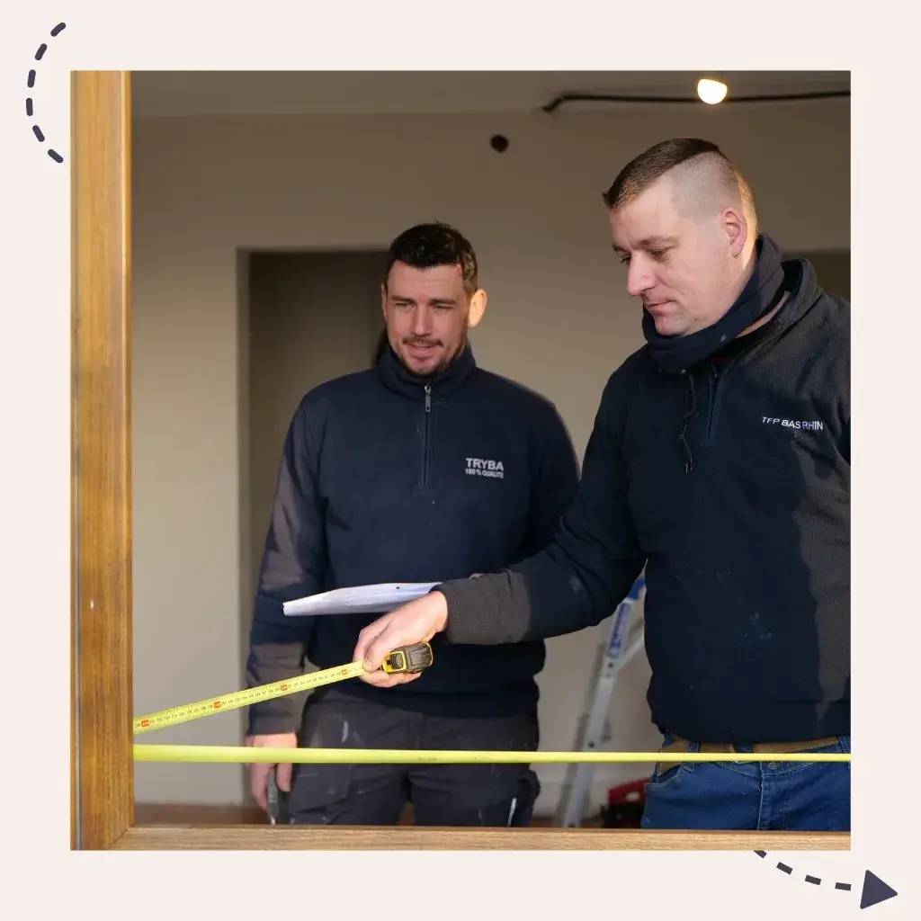 Deux hommes en tenue de travail mesurent un cadre en bois à l'aide d'un ruban à mesurer.