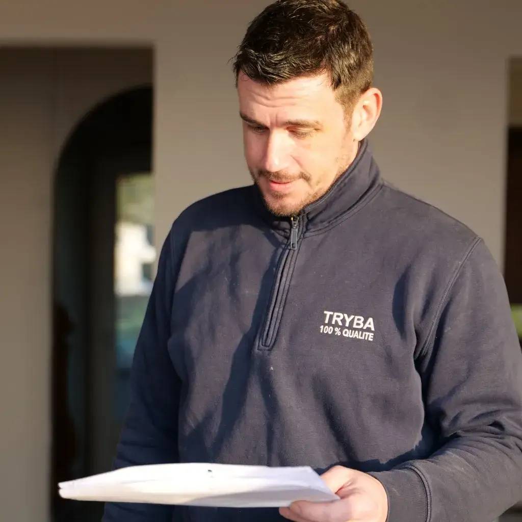 Un homme en pull bleu marqué "TRYBA 100% QUALITÉ" examine des documents à la lumière du jour.