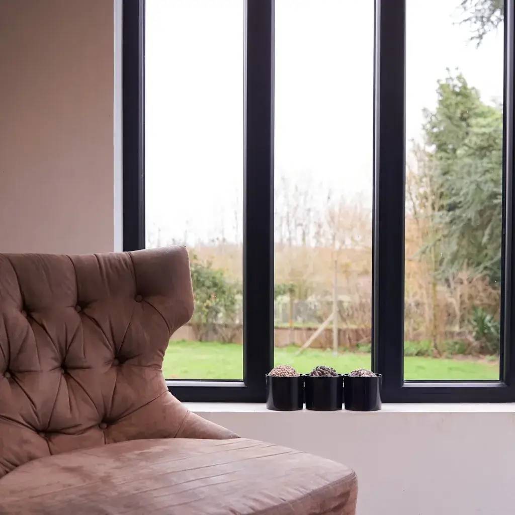 Un fauteuil en velours marron côtelé est placé près d'une fenêtre, offrant une vue sur un jardin verdoyant, avec trois petites plantes en