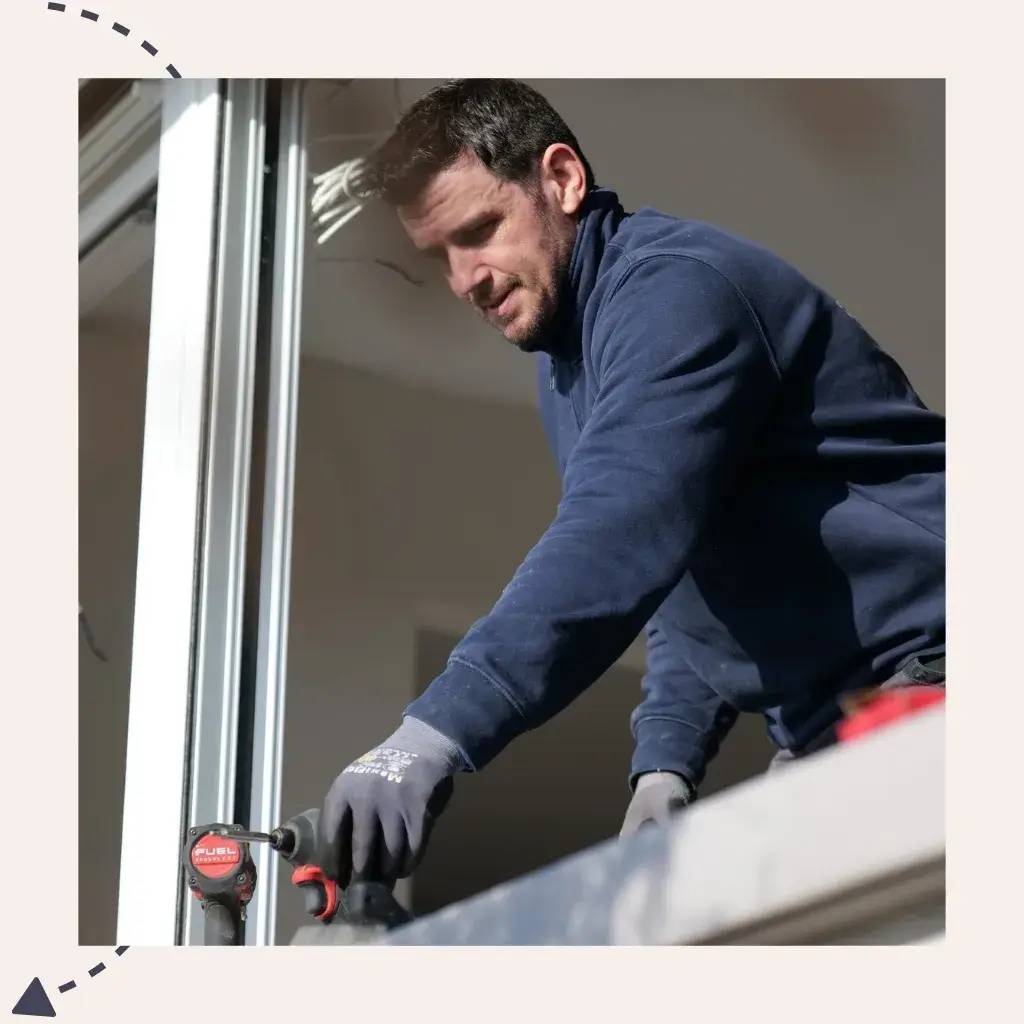 Un homme en tenue de travail installe une fenêtre en utilisant une perceuse électrique.