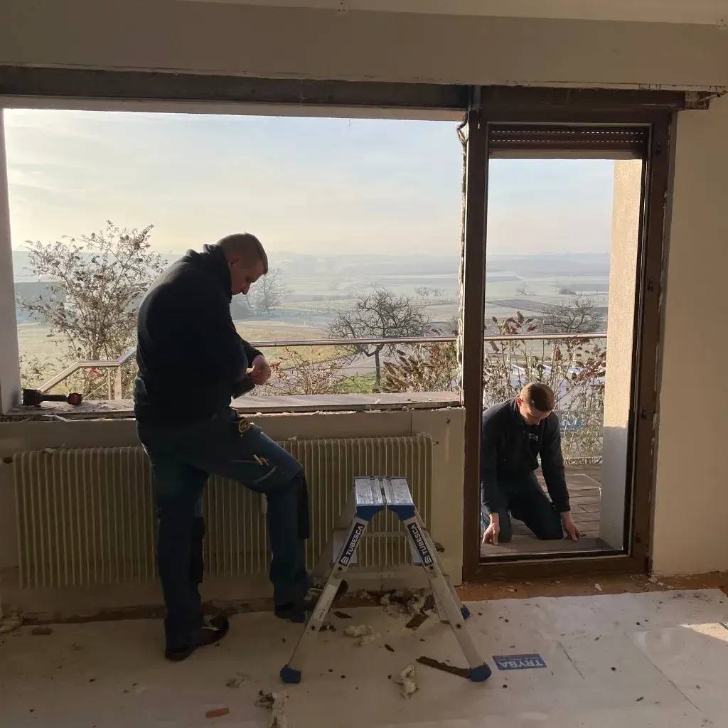 Deux personnes travaillent à l'installation d'une grande fenêtre dans une pièce lumineuse avec vue sur la campagne.