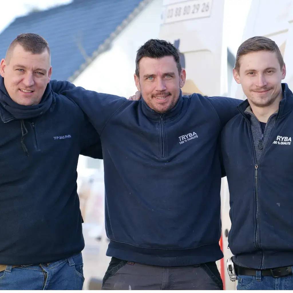 Trois hommes souriants en tenue de travail posent ensemble à l'extérieur, devant un bâtiment aux murs clairs.