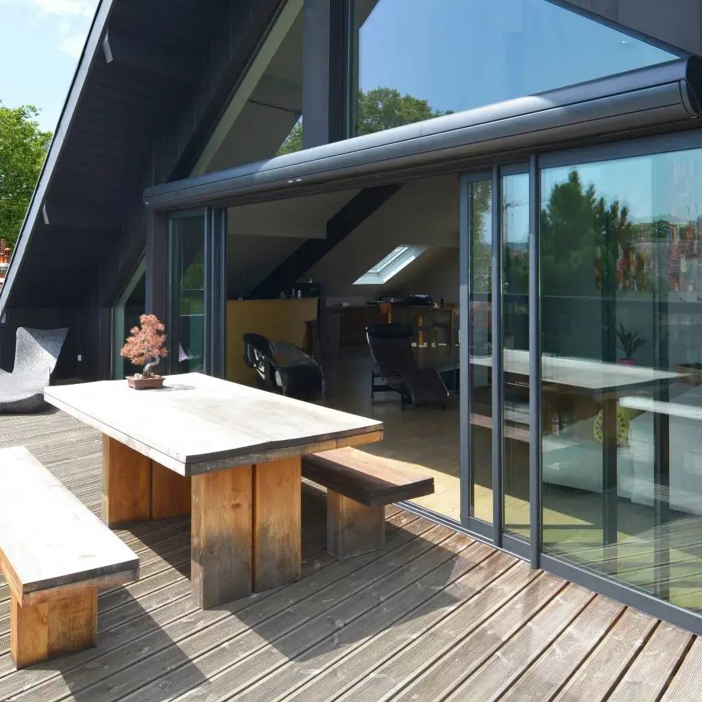 Une terrasse en bois avec une grande table rustique, adossée à une maison moderne aux grandes baies vitrées.