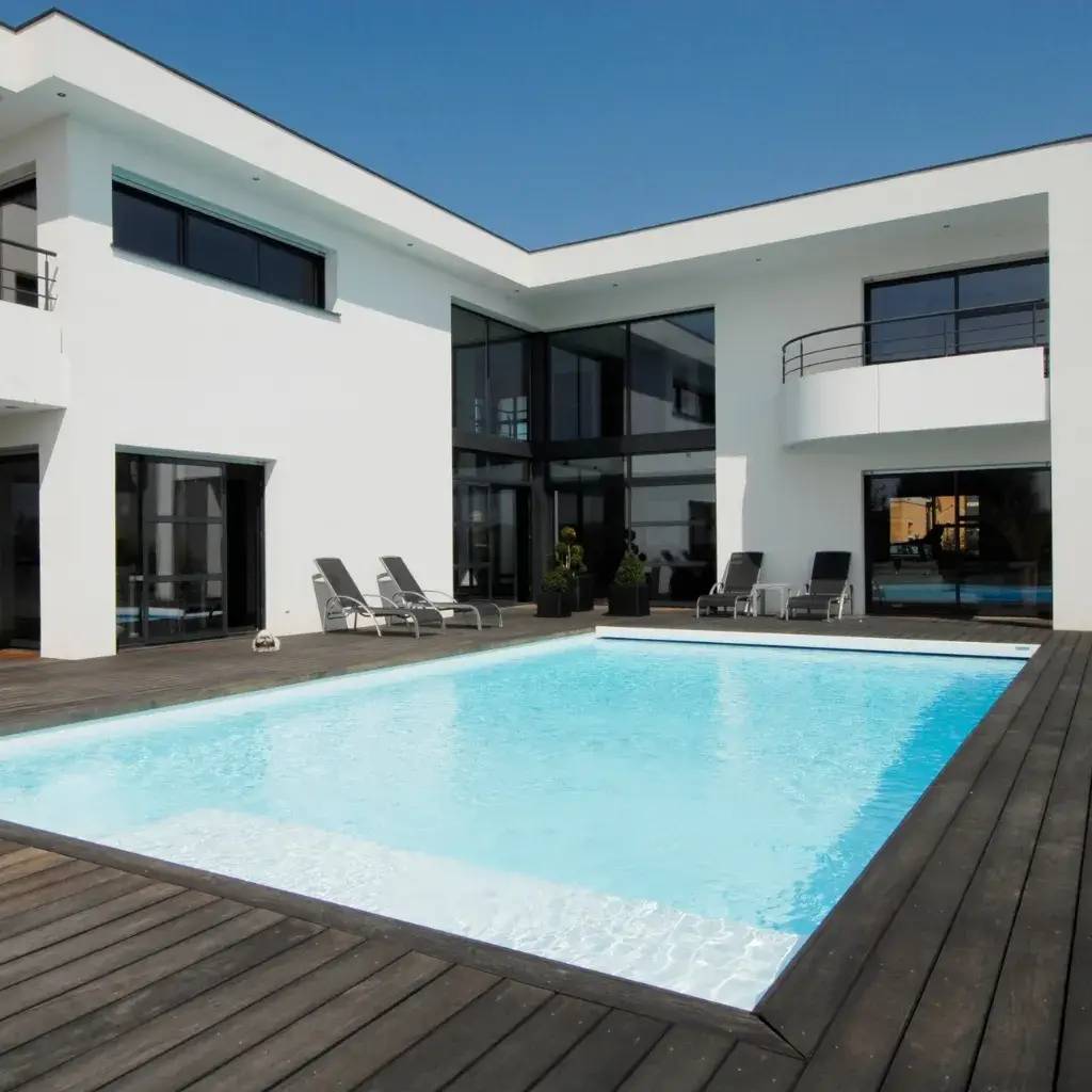 Une villa moderne avec une grande piscine entourée d'une terrasse en bois et équipée de chaises longues.