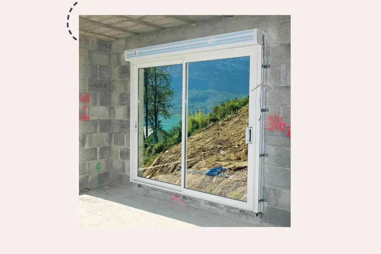 Une baie vitrée dans un mur en parpaings offre une vue sur un paysage vert et montagneux.