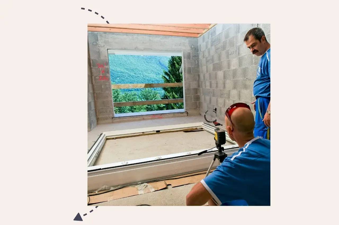 Des ouvriers en tenue bleue utilisent un équipement de mesure dans une pièce en construction avec une grande fenêtre ouverte sur un paysage verdoyant.
