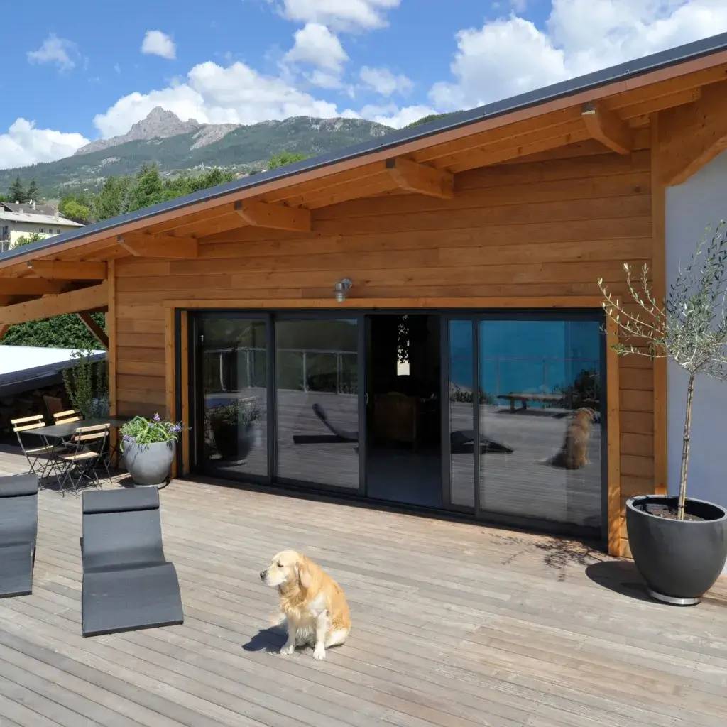 Une maison en bois avec une large terrasse, offrant une vue sur les montagnes et un chien assis paisiblement au soleil.