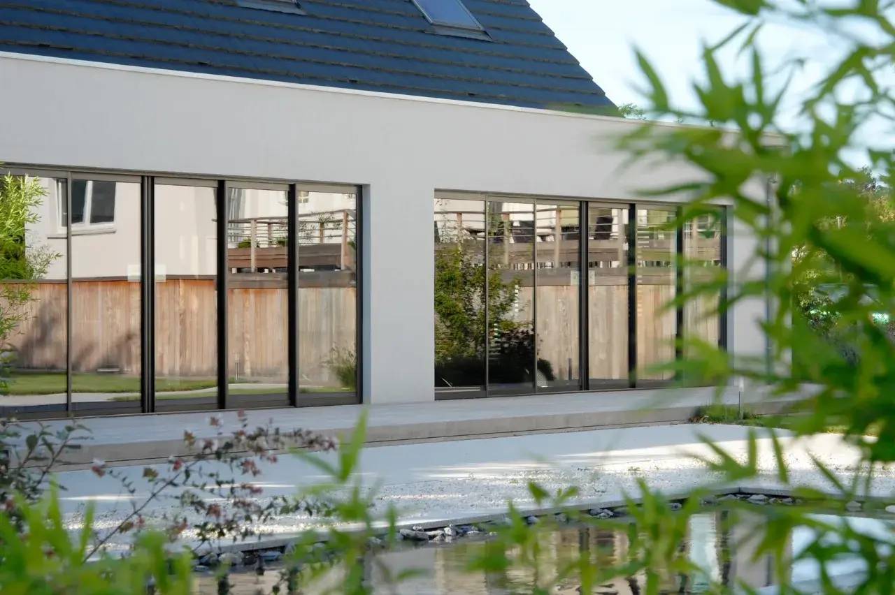 Vue d'une maison moderne avec de grandes fenêtres vitrées, entourée de verdure.