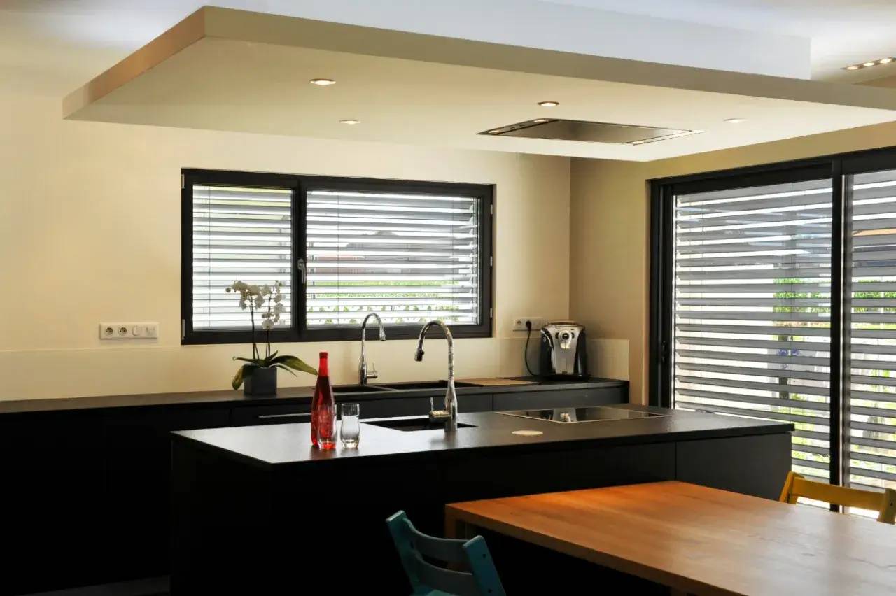 Cuisine moderne et lumineuse avec un îlot central noir, des fenêtres à persiennes et des éléments en acier inoxydable.