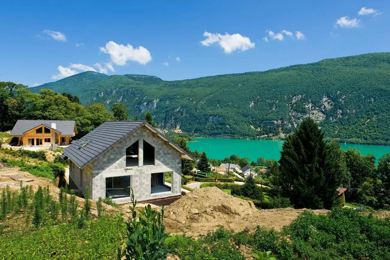 Une maison en construction se trouve au bord d'un lac aux eaux turquoise, entourée de collines verdoyantes et sous un ciel bleu.