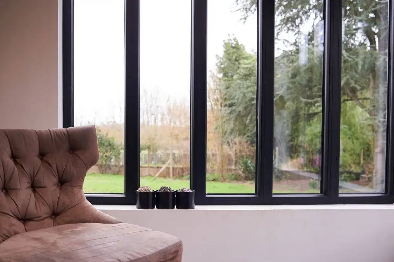 Un fauteuil beige confortable est placé près d'une grande baie vitrée donnant sur un jardin verdoyant, avec des petites plantes en pots sur le rebord