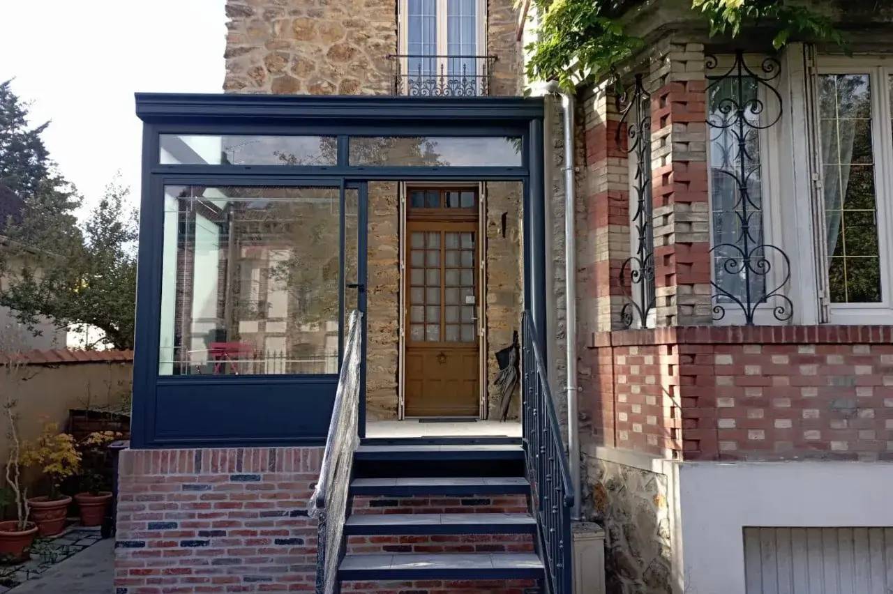 Entrée en verre moderne avec escalier en briques et porte en bois, intégrée à une façade en pierre et briques.