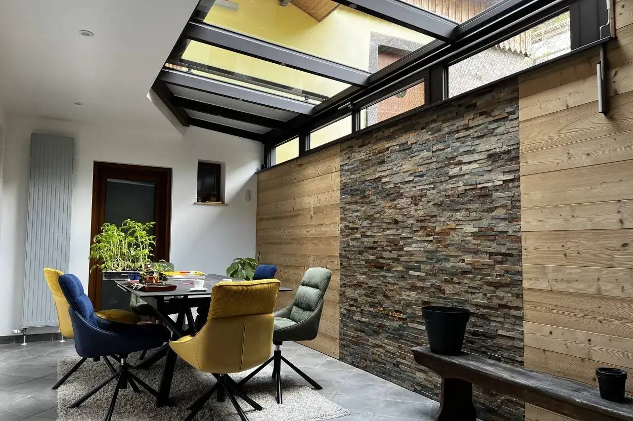 Un espace de vie moderne et lumineux avec un toit en verre, des chaises colorées autour d'une table, et un mur en pierre naturelle.