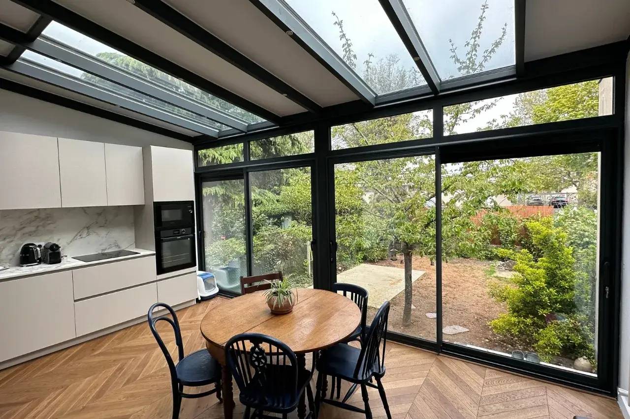 Cette cuisine lumineuse, dotée de grandes baies vitrées et d'un plafond en verre, offre une vue panoramique sur un jardin verdoyant