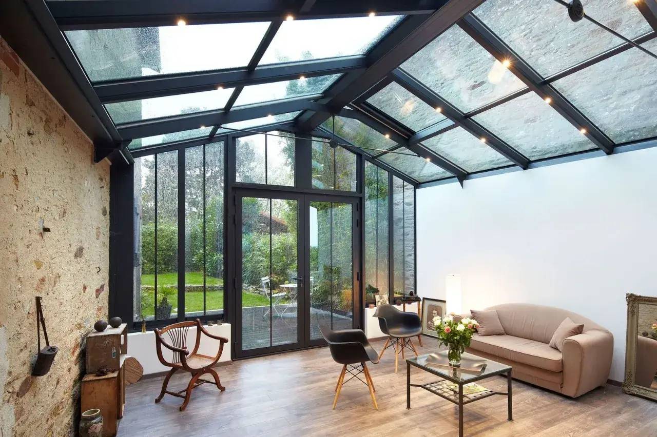 Une véranda moderne avec un toit en verre, offrant une vue dégagée sur le jardin, abrite un espace de détente élégant avec des fauteuils